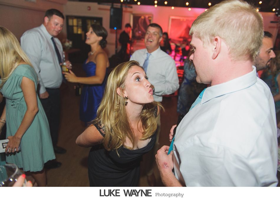 Woman puckering lips toward a man in a white shirt at a party. Other guests laugh in the background.