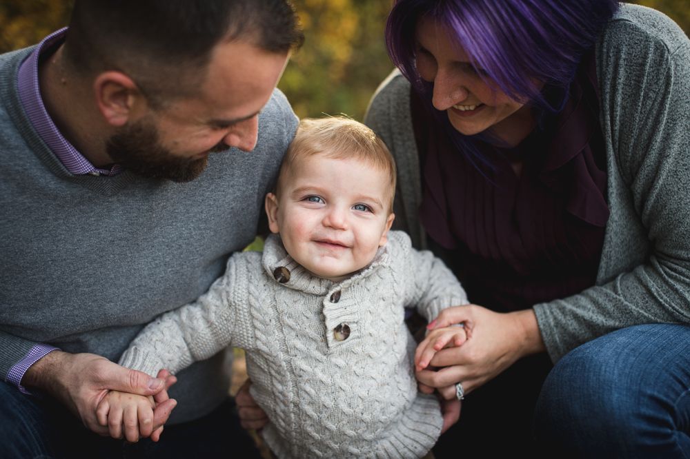 Parents holding a smiling toddler in a cozy sweater outdoors.