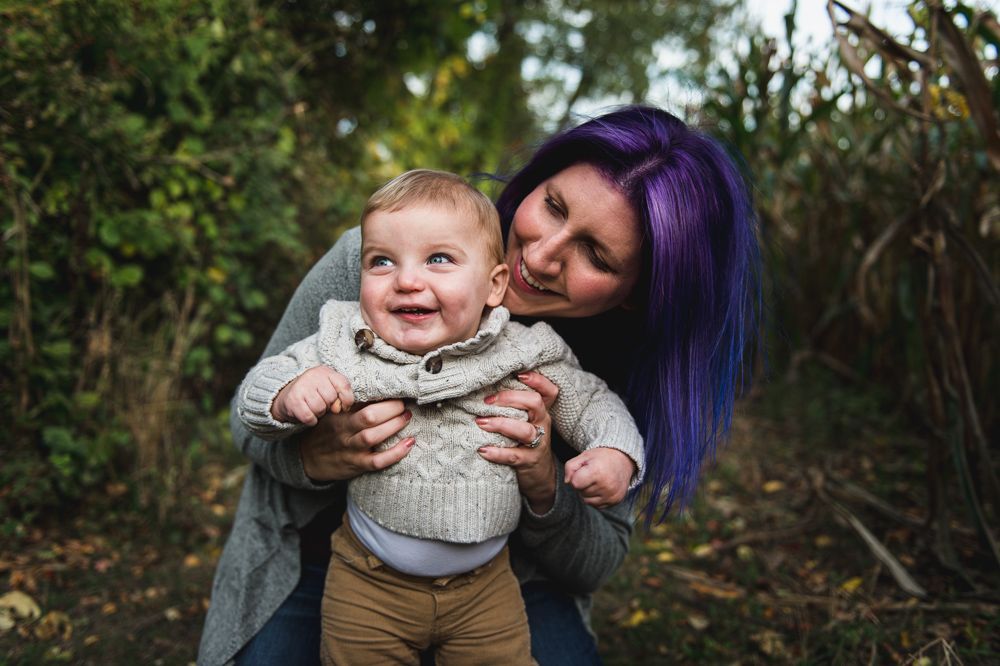 Woman with purple hair smiling at a smiling baby, holding him up. They are outdoors, surrounded by foliage.