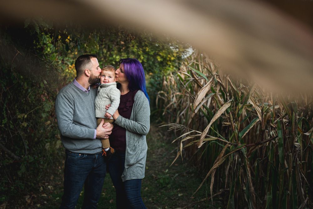 Family of three in a cornfield, mother kissing child's head, father holding child, autumn foliage.