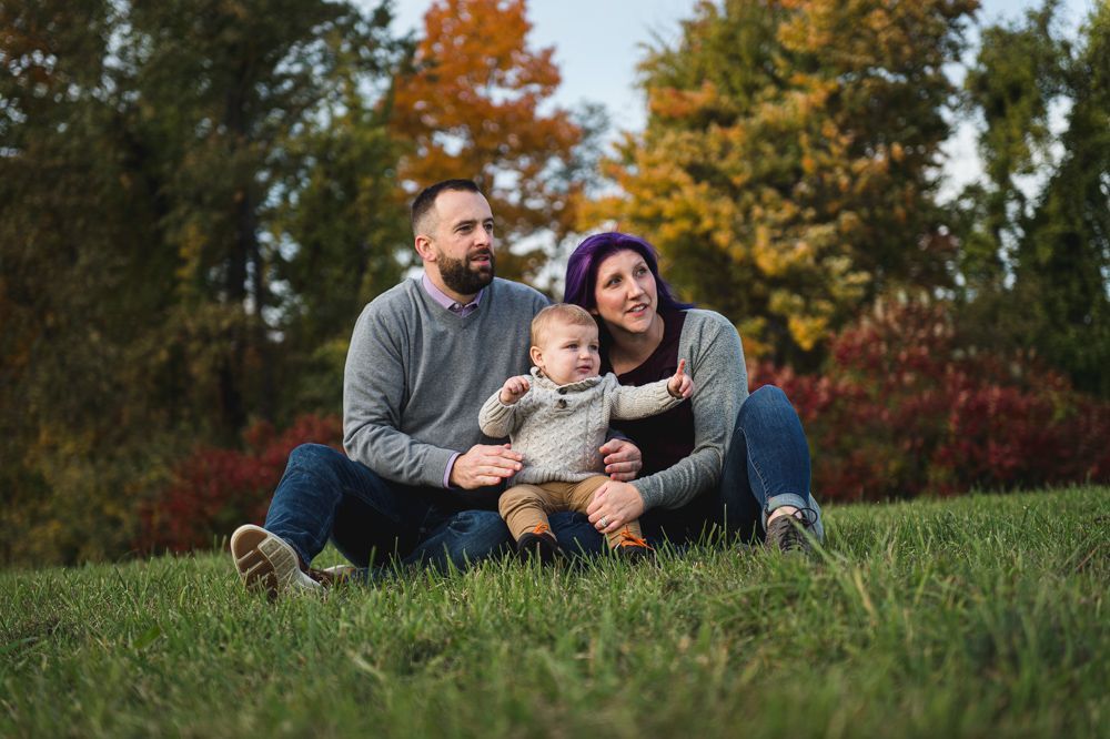 Family of three sitting on grass, looking upwards. Autumn foliage in background.