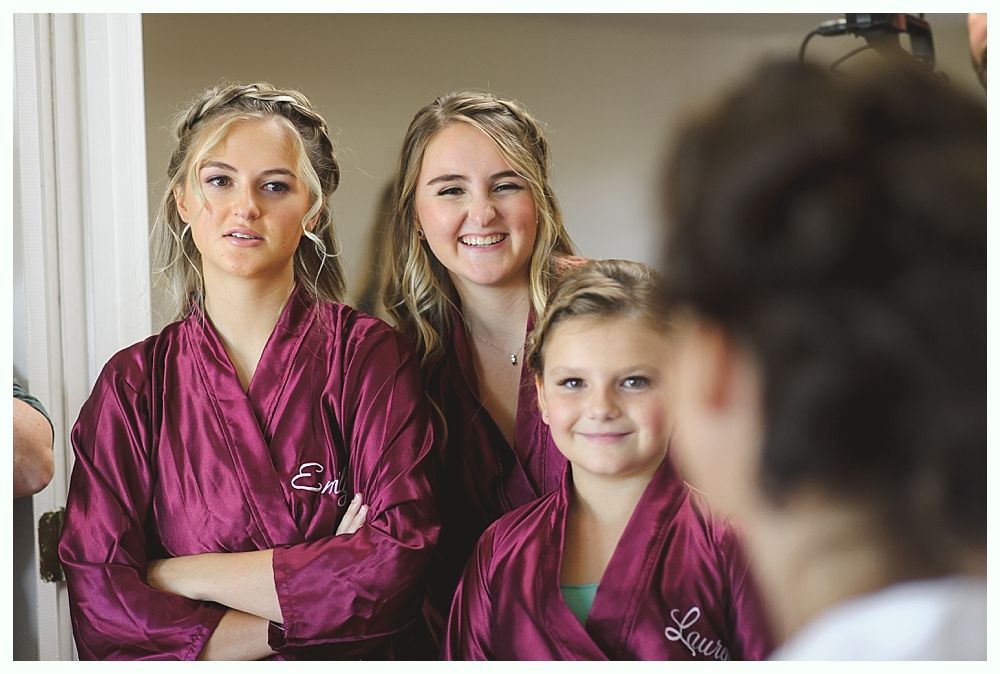 Three women in matching burgundy robes, two smiling, one with arms crossed, in a room with a mirror.