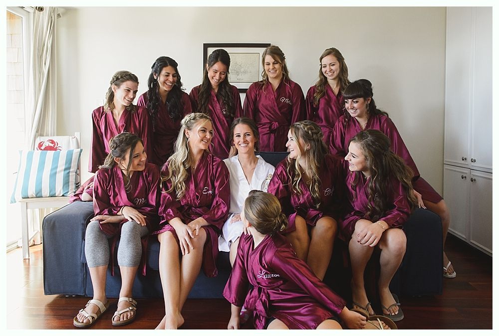 Bridesmaids in burgundy robes with bride in white robe, sitting on a couch, smiling indoors.