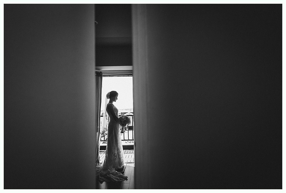 Bride in silhouette standing between walls, holding bouquet, looking out a window. Black and white.