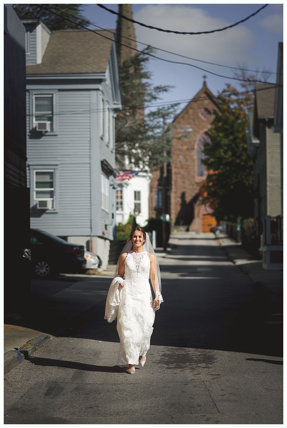 Woman in white wedding dress walks down a street toward a church on a sunny day.