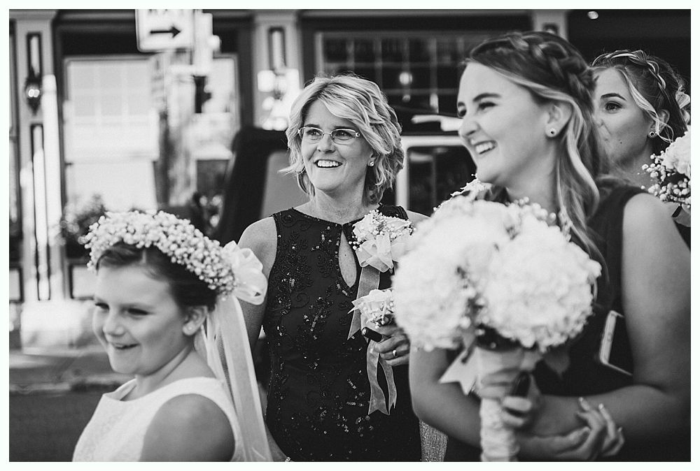 Wedding party smiling, holding bouquets outside a building. Black and white photo.