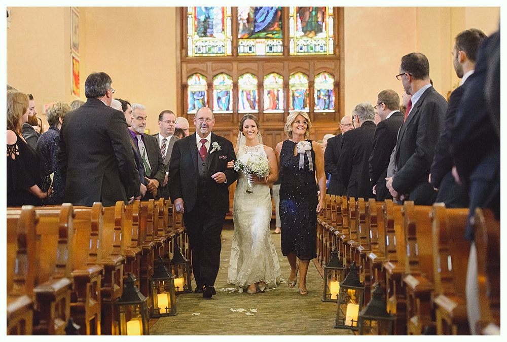 Bride walks down aisle with her father, mother following, towards stained glass windows in church.