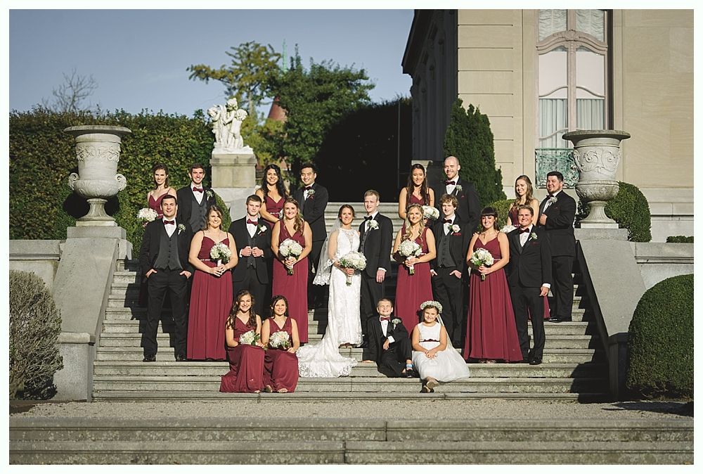 Wedding party on steps, bride in white dress, bridesmaids in red, groomsmen in black, outdoor mansion setting.