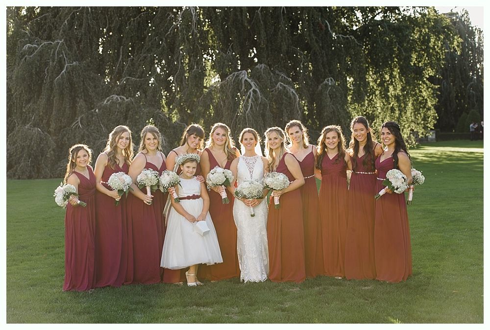 Bride with bridesmaids in burgundy dresses and flower girl, outdoors in front of trees, holding bouquets.