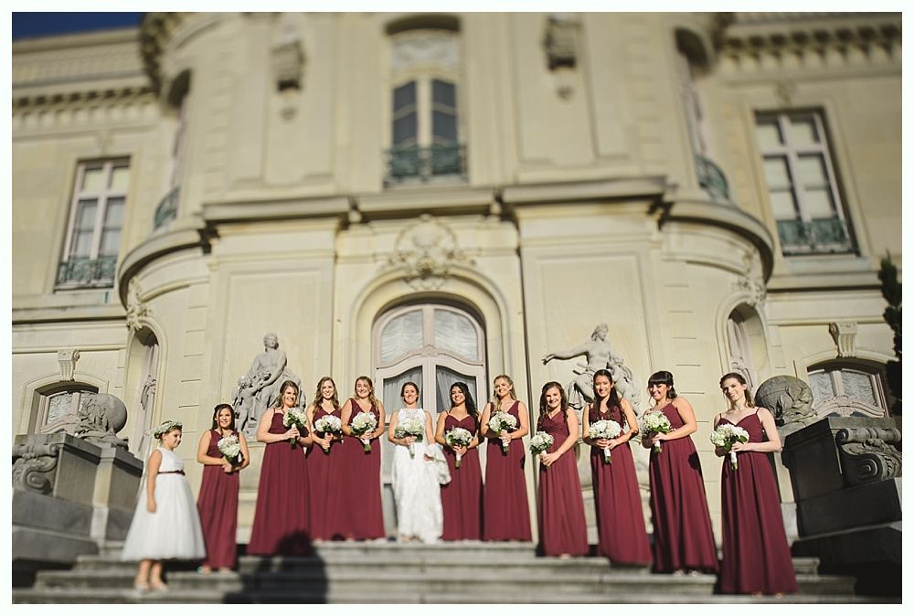 Bridesmaids in burgundy dresses and a bride in white pose on steps in front of a large building.