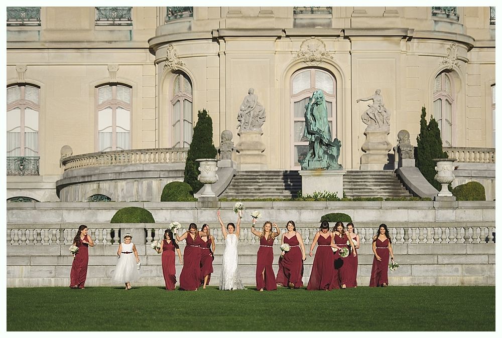 Bridesmaids in burgundy and the bride in white pose on a lawn in front of a grand building.