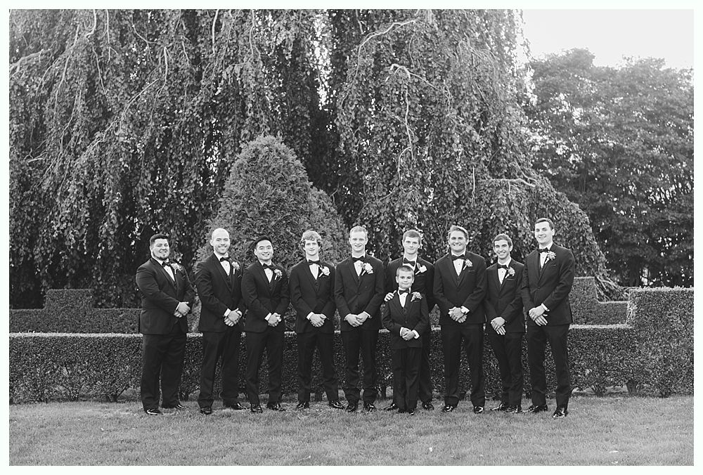 Group of men in tuxedos posing outdoors in front of leafy trees.