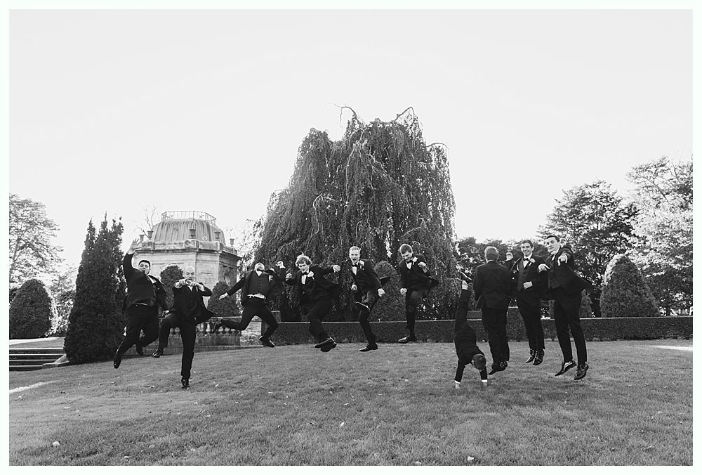 Men in tuxedos jumping in a grassy outdoor setting, a building and large tree in the background.