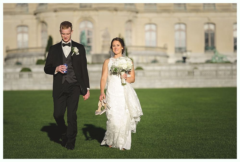 Couple walking on a green lawn in front of a large mansion, bride holding bouquet and shoes, groom holding a drink.