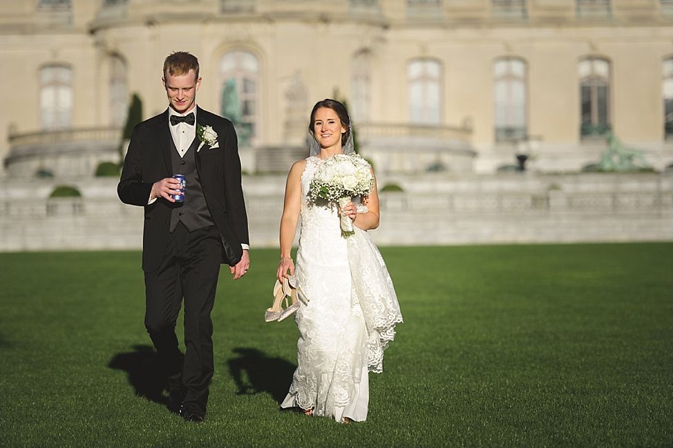 Bride and groom walking on lawn in front of a large building. The bride carries her shoes and bouquet. Groom holds a drink.