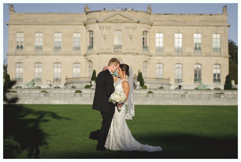 Bride and groom embrace in front of a grand mansion. Sunny day, green lawn.