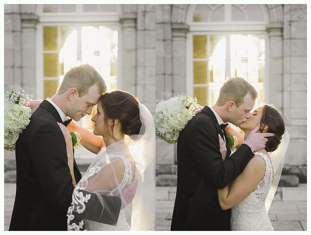 Bride and groom in formal wear embrace and kiss, with a bouquet, in front of a building with columns and large windows.