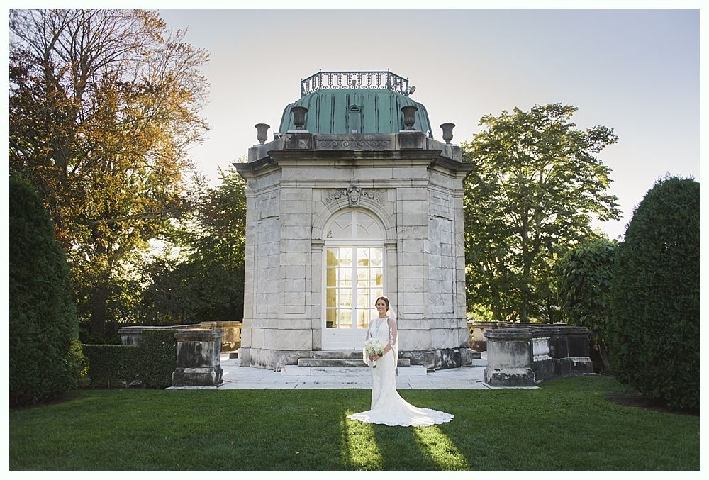 Bride in a white gown stands in front of a stone pavilion with a green dome; sunny outdoor setting.