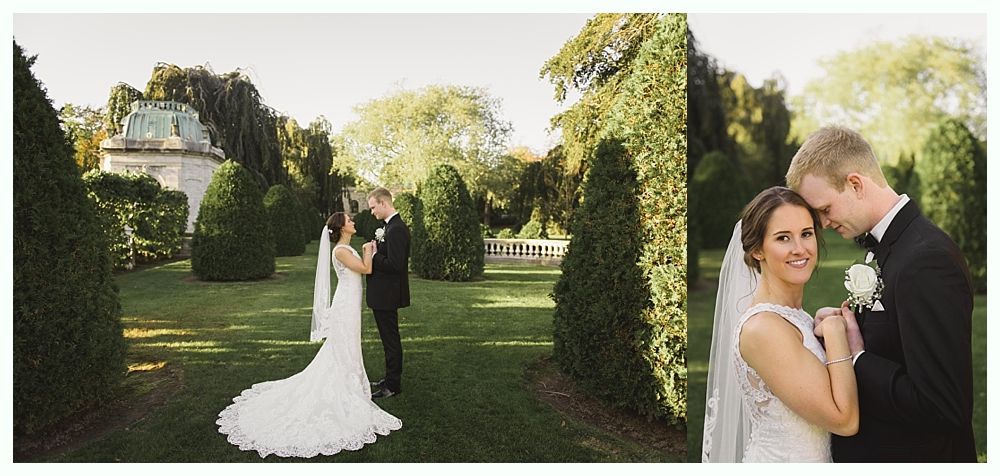 Newlyweds in wedding attire embrace in a garden with greenery and architecture.
