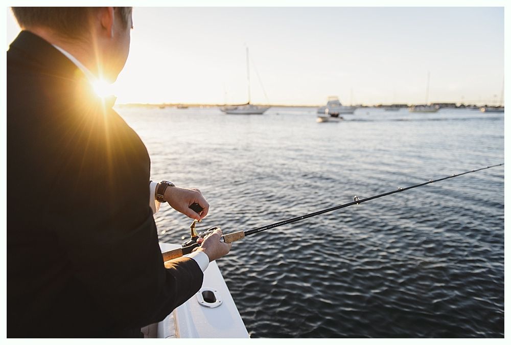 Man in suit fishing off a boat at sunset, sailboats in background.