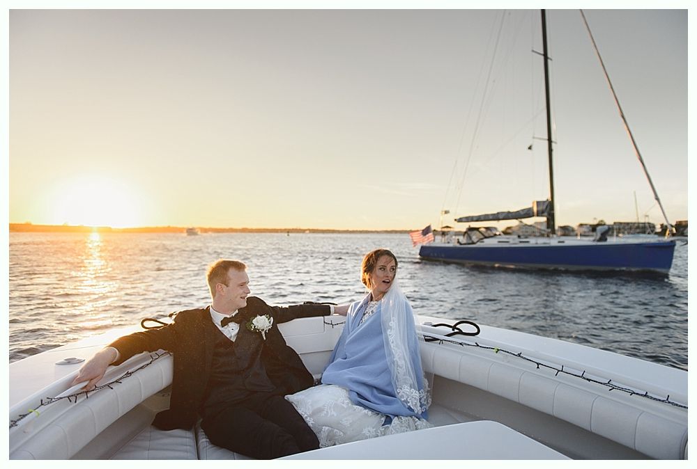 Newlyweds in a small boat on water at sunset, sailboat in background. Bride wrapped in a blue shawl.