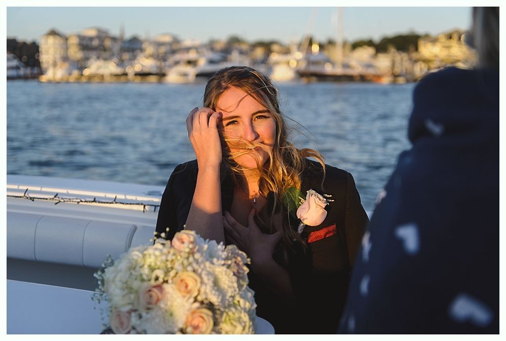 Woman on a boat, holding flowers. She touches her hair; sunset illuminates her face. Water and buildings in background.