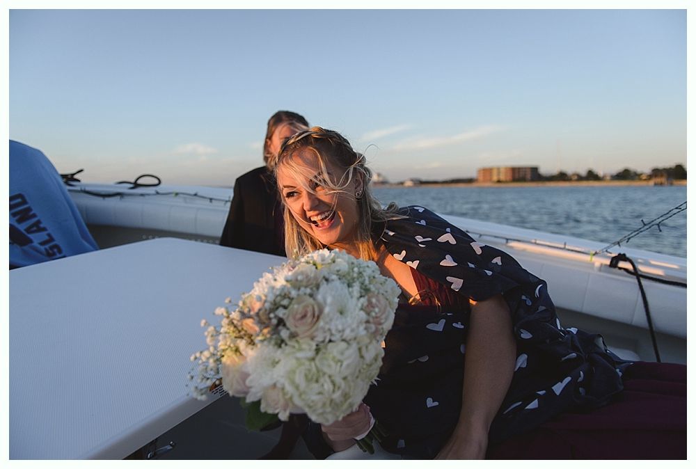 Woman on a boat laughs, holding a bouquet. Behind her, a person in a black suit. Sunny, outdoor setting.