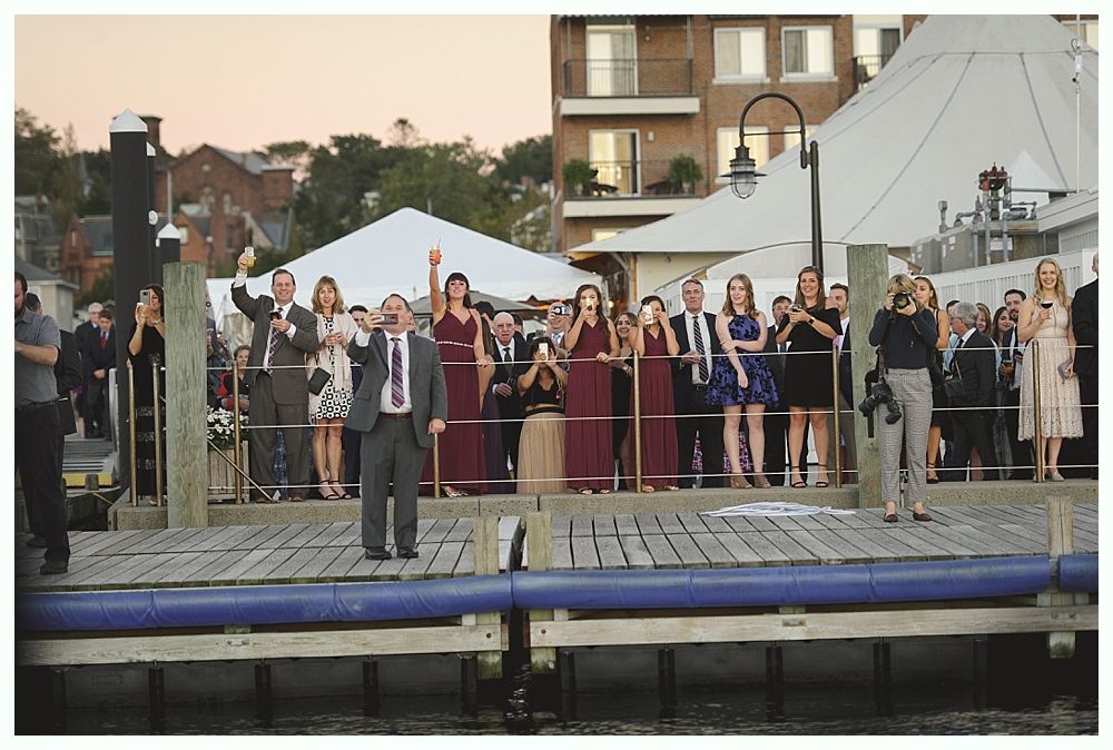 People gathered on a dock, raising their arms and cheering. Buildings and a tent are in the background, near water.