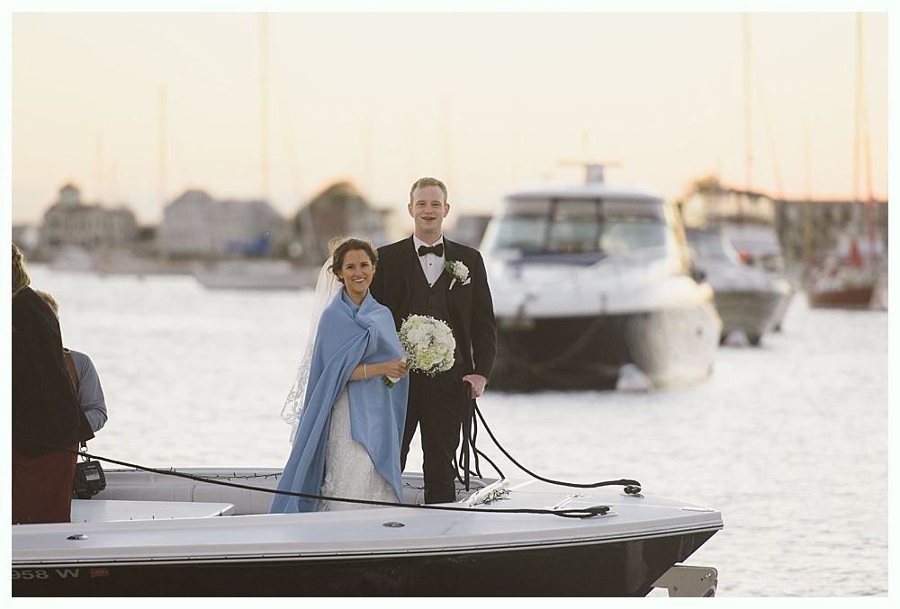 Newlyweds on boat by water. Bride in blue shawl, groom in suit. Boats and harbor in the background.
