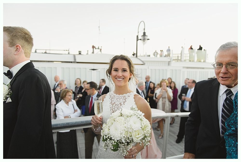 Bride holding flowers and champagne smiles at the camera on a pier, with wedding guests behind her.