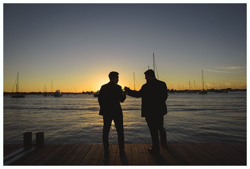 Two silhouetted people on a dock at sunset, near boats.