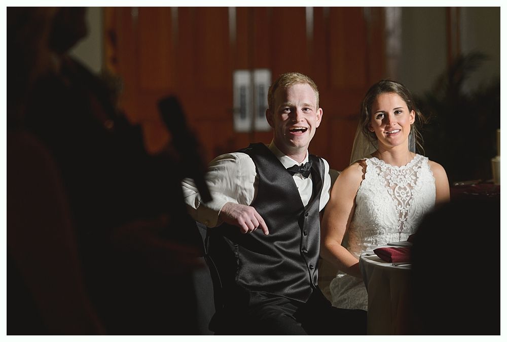 Newlyweds at a reception: Smiling groom in vest, bride in lace dress, dark setting.