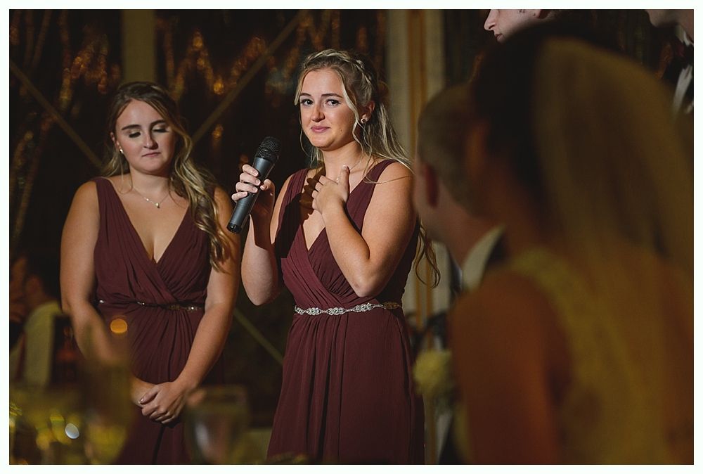 Woman in burgundy dress gives a speech at a wedding, appearing emotional. Another woman stands beside her.