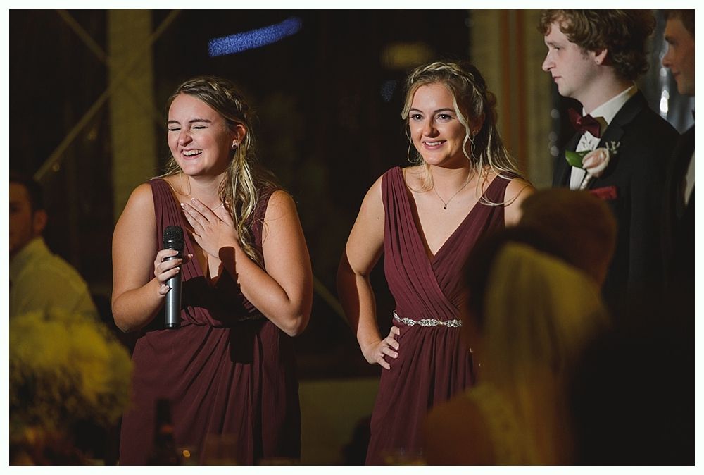 Two women in burgundy dresses giving a speech at a wedding reception. One holds a microphone, laughing.