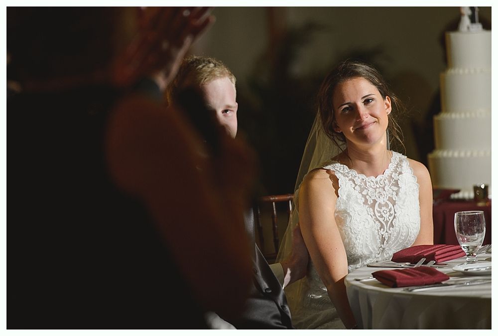 Bride and groom listening to a speech at wedding reception; cake in background.
