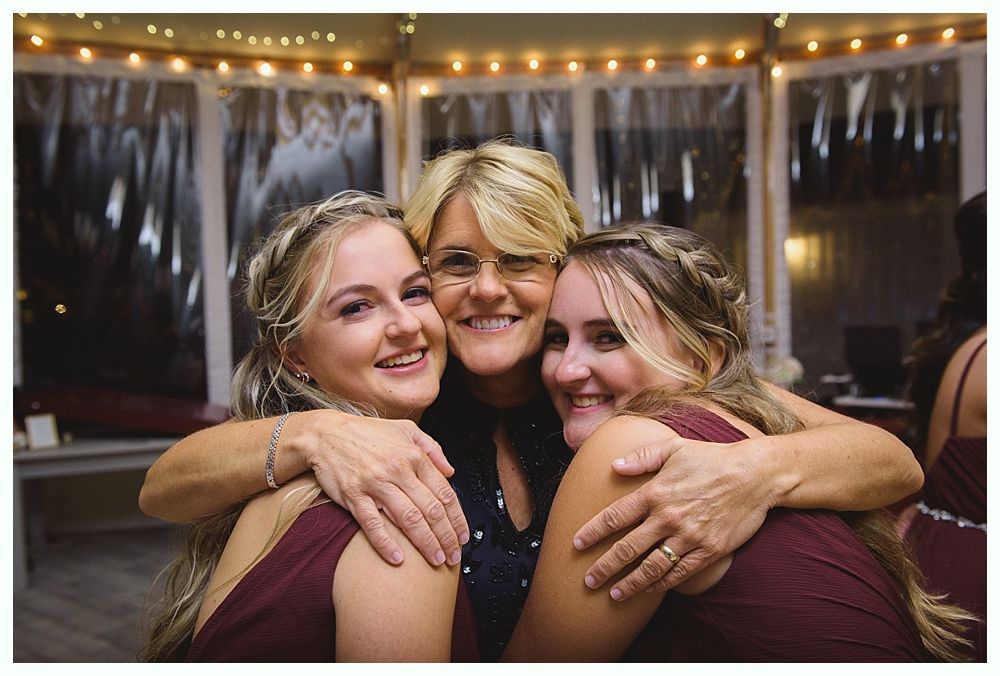 Woman hugging two people in maroon dresses, smiling. Lit by string lights inside a tent.