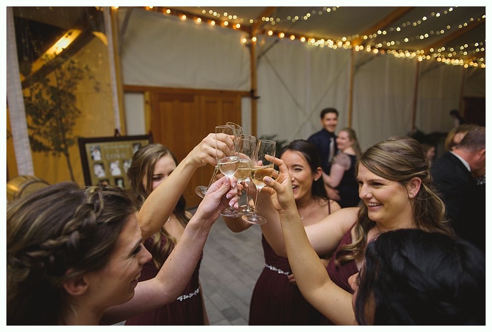 Bridesmaids toasting with champagne flutes at a wedding reception. Lights overhead; wooden beams and a framed photo are visible.