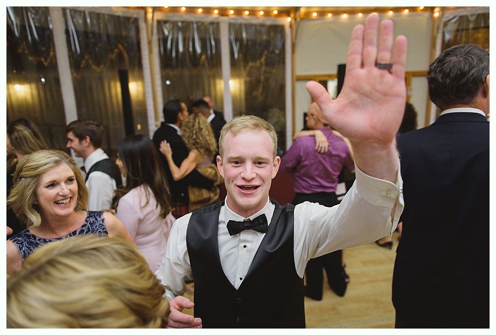 Man in a vest and bow tie raises his hand at a party, smiling. People dance in the background.