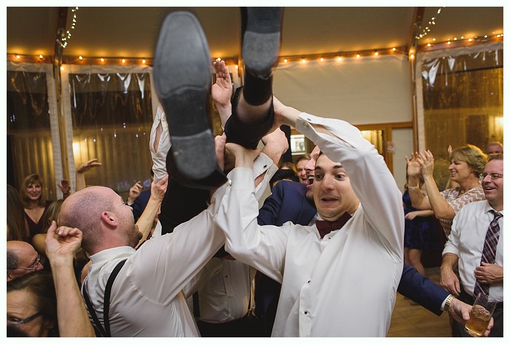 Men lifting a person in dress shoes during a celebration inside a room with string lights.