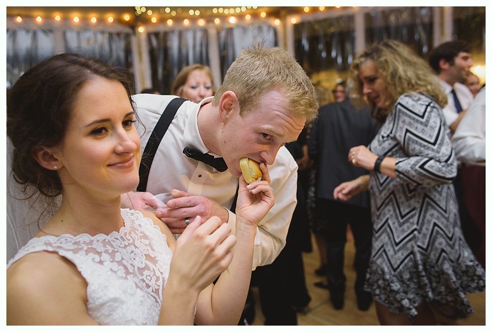 Newlyweds at wedding reception. Groom eating food with bride looking on. People dancing in background.