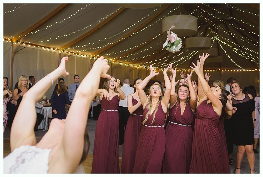 Bride throwing bouquet toward a group of women in burgundy dresses. Reception hall with string lights.