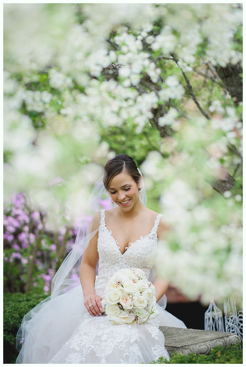 Bride in white dress holding bouquet, smiles near flowering trees.