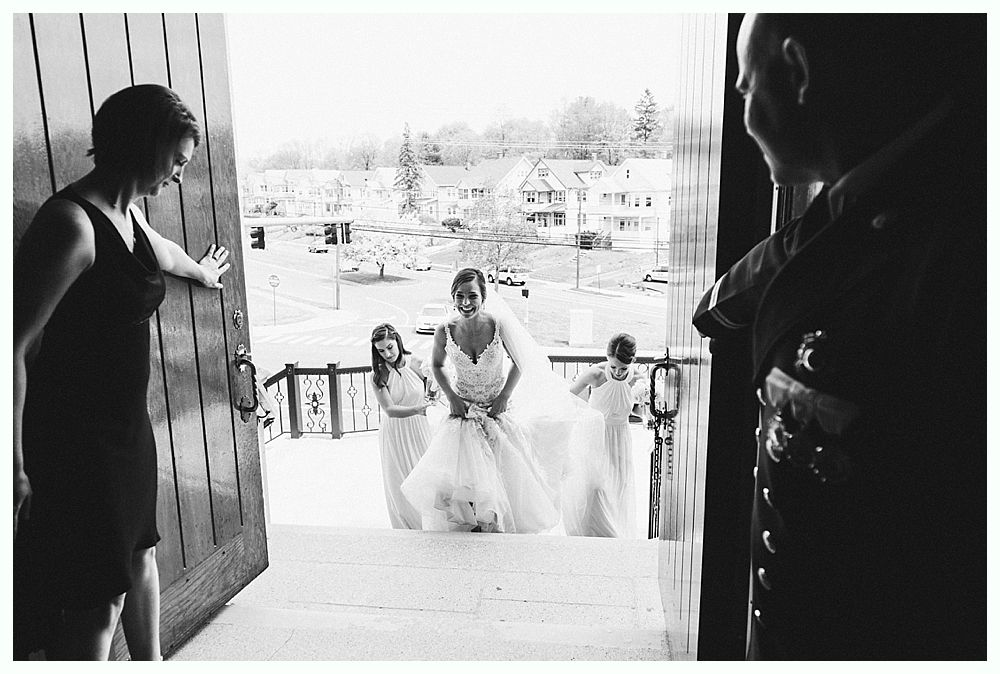 Bride at church entrance, awaiting ceremony. Bridesmaids in white gowns and a woman in a black dress by the doors.