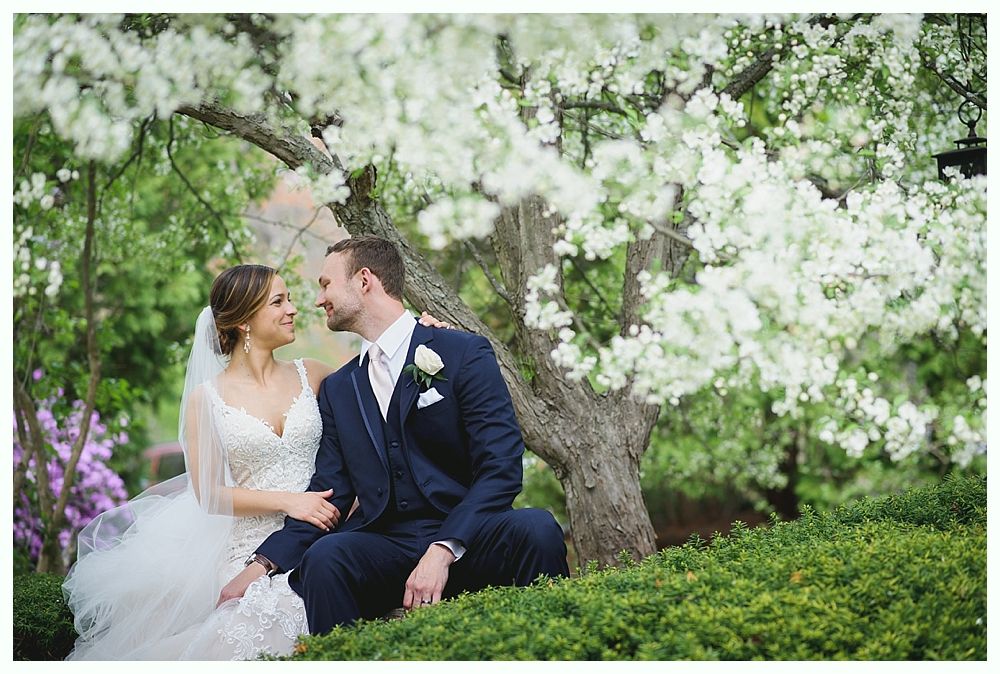 Newlyweds sit under a flowering tree, looking at each other; bride in a white dress, groom in a blue suit.