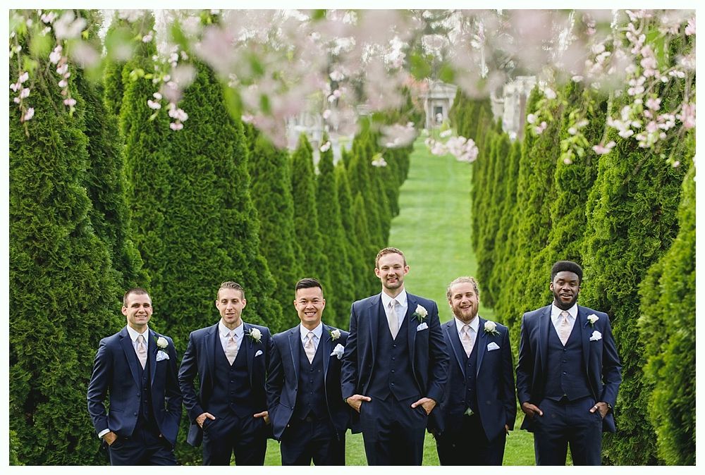 Men in navy suits stand outdoors, tall green trees behind them.