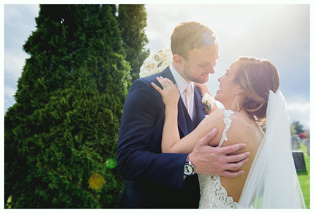 Bride and groom embracing, looking at each other. Sunlight, green tree, and cloudy sky in background.