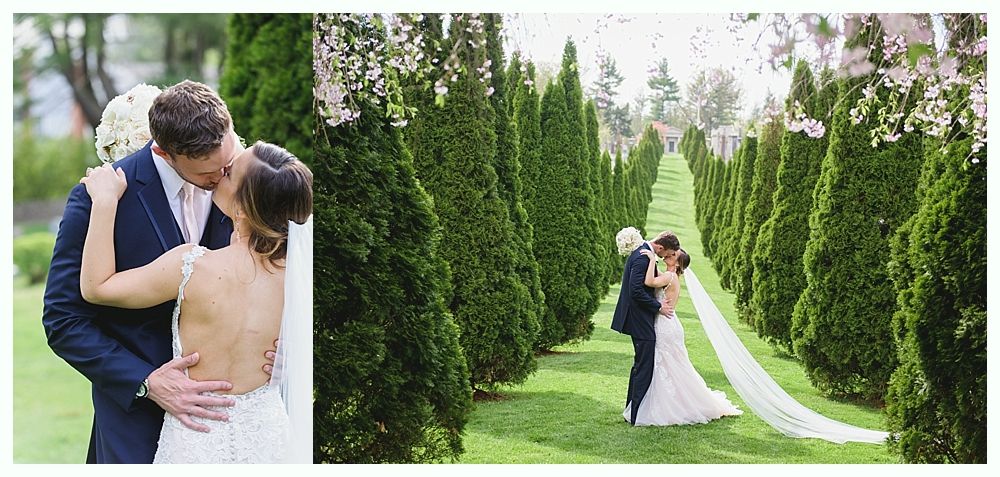 Newlyweds share a kiss in a garden lined with tall green trees. The bride's veil trails behind them.