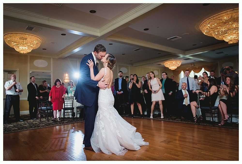 Couple kissing during their first dance at a wedding reception; guests watch, chandeliers illuminate the room.