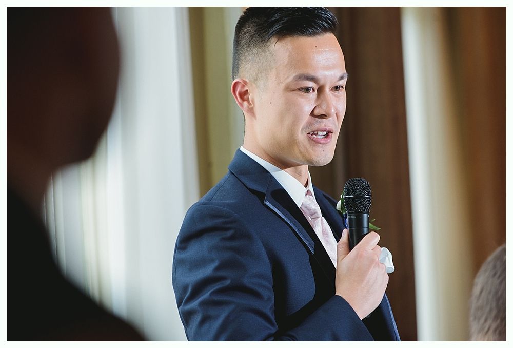 Man in navy suit speaks into a microphone, indoors, with a pink tie, and a blurred person in the foreground.
