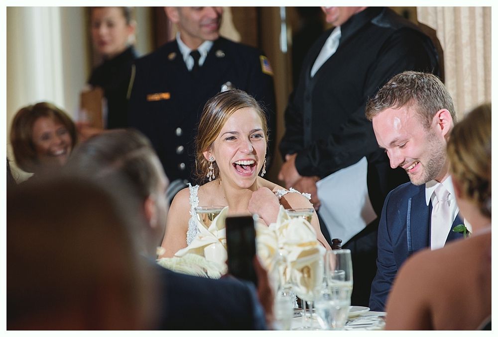 Bride laughing at a wedding reception, seated at a table with a man, and other guests nearby.